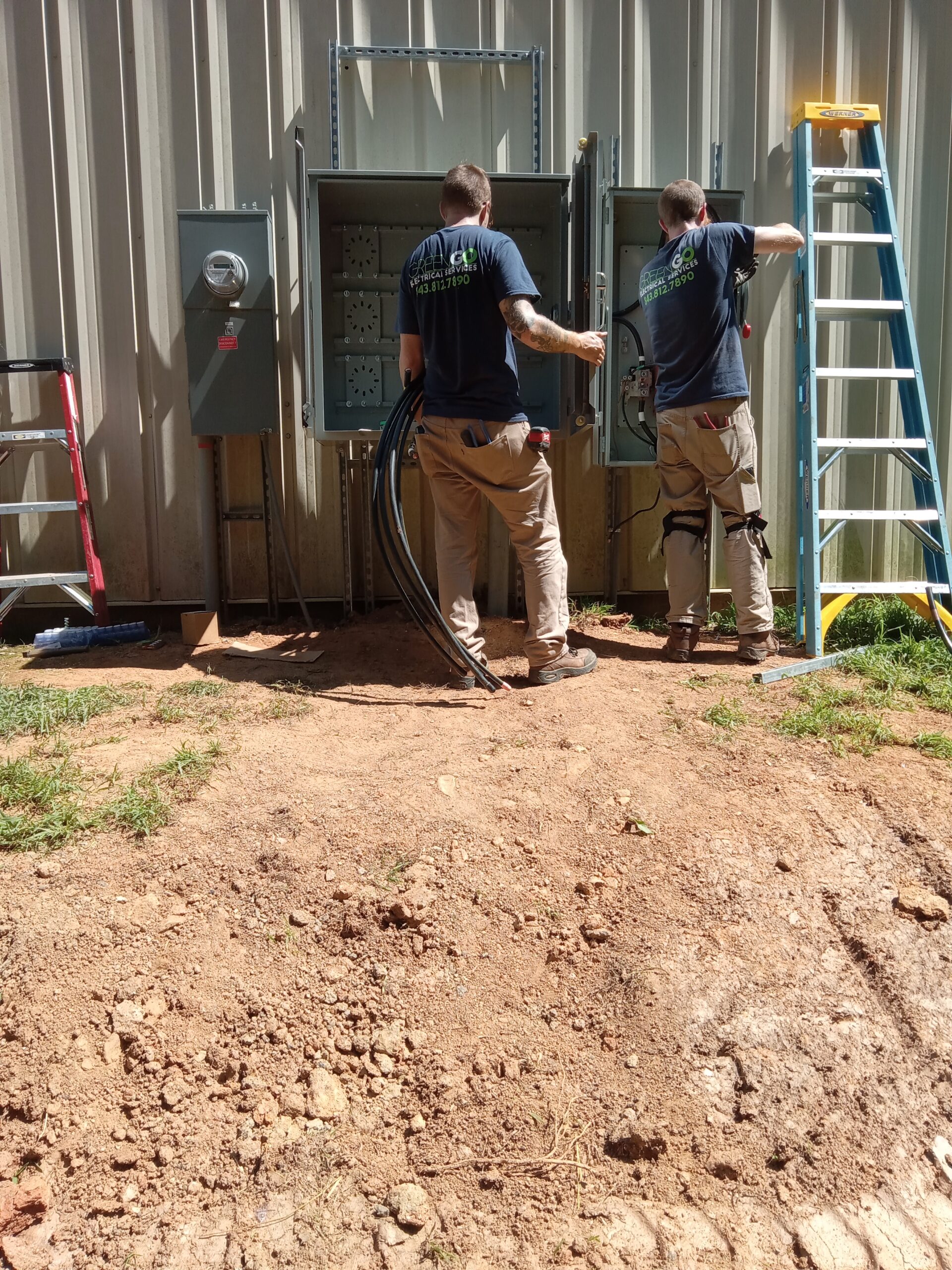 Two workers in blue shirts and khaki pants install electrical equipment outdoors next to a building. They are using ladders and handling cables.