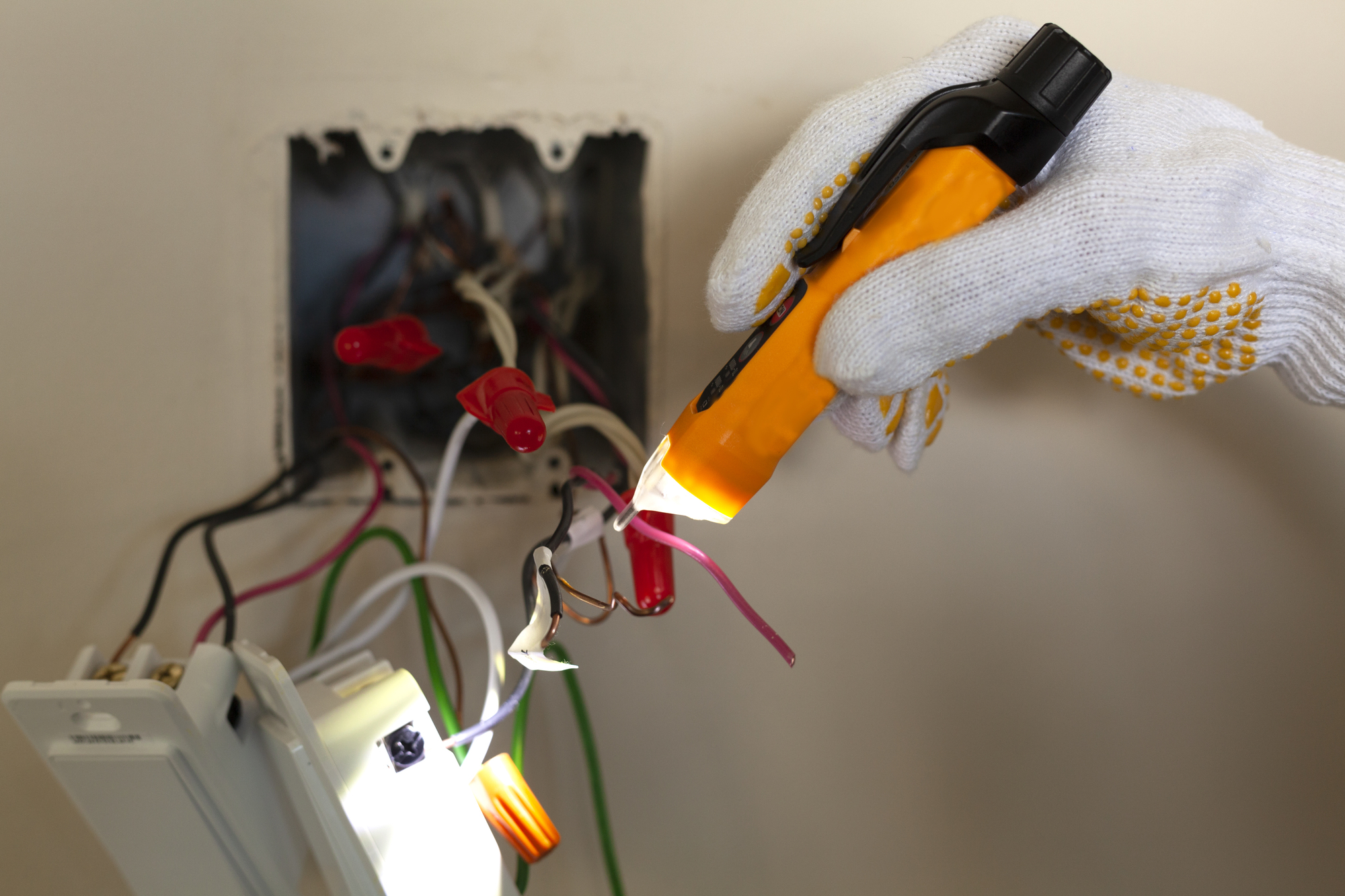 A person wearing gloves uses a voltage tester on exposed electrical wires next to an open wall outlet.