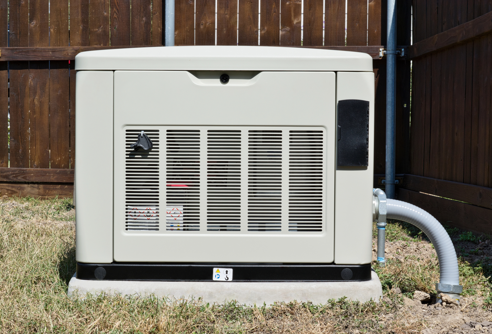 A beige backup generator is installed on a concrete slab next to a wooden fence. It has a vented front panel and a cable connected on the side.