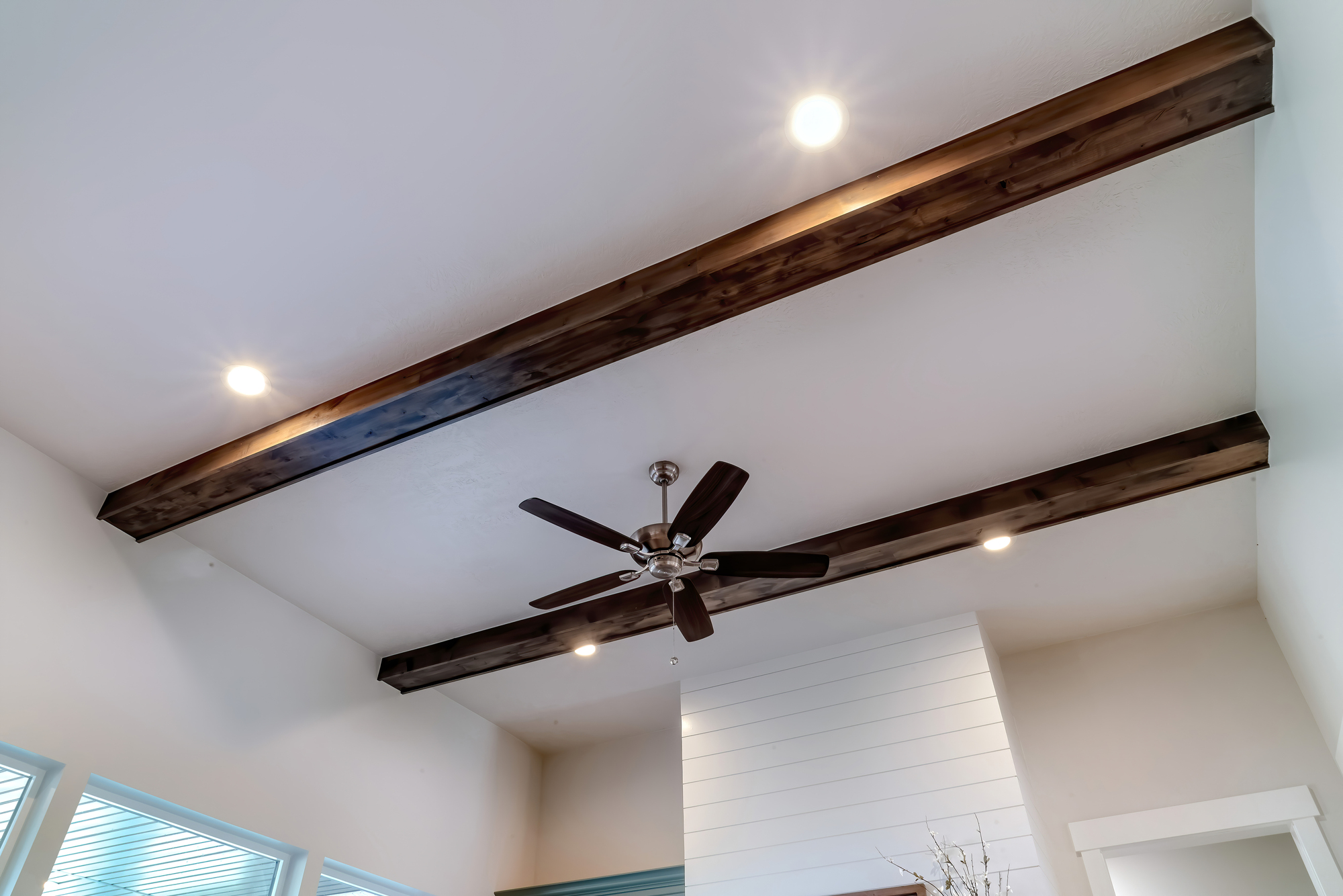 Ceiling with two wooden beams, a dark brown ceiling fan, and recessed lights in a modern room with white walls and windows.
